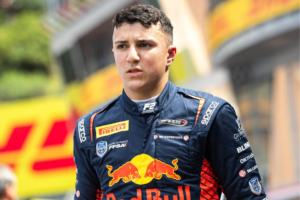 O piloto de automobilismo Isack Hadjar em primeiro plano, vestindo um macacão de corrida azul-escuro com o logotipo em destaque da Red Bull e patrocinadores como Campos Racing, Pirelli e Sparco. Ele é um homem jovem, de cabelos curtos e escuros, olhar concentrado direcionado levemente para a esquerda. Ao fundo, fora de foco, vê-se a estrutura de um autódromo com detalhes em amarelo e vermelho.
