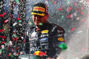 O piloto de Fórmula 1 Max Verstappen sorri no pódio enquanto é coberto por spray de espumante e confetes coloridos em comemoração. Ele veste o macacão escuro da Red Bull Racing com diversos patrocinadores em destaque e segura a garrafa de espumante na altura do peito, usando um boné preto da Pirelli com detalhes dourados.