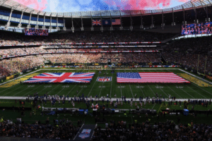 Vista panorâmica do Tottenham Hotspur Stadium lotado durante um jogo da NFL, com bandeiras gigantes do Reino Unido e dos Estados Unidos estendidas sobre o gramado antes da partida, arquibancadas cheias de torcedores e céu ao entardecer com nuvens rosadas ao fundo.