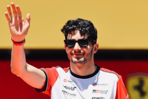 Esteban Ocon, piloto da Haas, acena com a mão direita e sorri de leve. Ele usa óculos escuros e o uniforme branco e vermelho da equipe, com logos da Haas, MoneyGram e Toyota Gazoo Racing. Ao fundo, há uma parede amarela e uma estrutura vermelha com parte de um emblema do cavalo rampante da Ferrari.