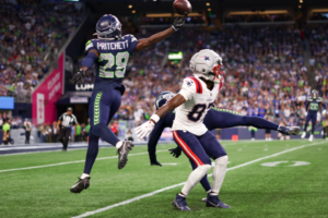 Foto de ação de um jogo da NFL entre o Seattle Seahawks e o New England Patriots. Neemias Pritchett, número 29 do Seahawks, é pego pulando alto no ar para pegar um passe, esticando o braço para cima para a bola. Um jogador do Patriots vestindo a camisa 84 está pulando à frente dele, com os braços estendidos, tentando contestar a recepção. A cena ocorre em um campo de grama verde com um grande estádio lotado ao fundo.