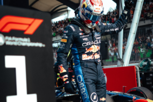 Max Verstappen com um macacão azul-marinho da Red Bull Racing e capacete com detalhes em vermelho e amarelo sai do cockpit do carro, com um braço erguido em comemoração. À esquerda, uma placa preta com o logotipo da F1 e o número 1 indica a primeira posição. Ao fundo, o público aparece nas arquibancadas