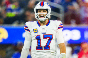 Quarterback do Buffalo Bills em close, de capacete branco com detalhes em vermelho e azul, gritando durante uma partida. Ele usa a camisa branca número 17 com listras azuis e vermelhas nas mangas e o patch “C” de capitão no peito. Ao fundo, o estádio aparece desfocado, com torcedores e painéis azuis.