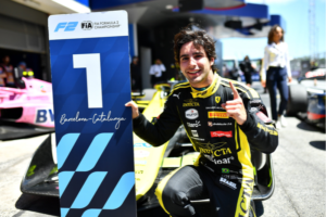 Rafael Câmara sorridente de macacão preto e amarelo, com logotipos de patrocinadores, agachado ao lado de um carro de corrida amarelo. Ele faz um sinal de positivo com a mão direita e apoia a outra mão em uma placa azul com o número 1 e a inscrição “Barcelona-Catalunya”, do campeonato FIA Fórmula 2. Ao fundo, em um dia ensolarado, aparecem um carro rosa e uma mulher de camisa branca caminhando.